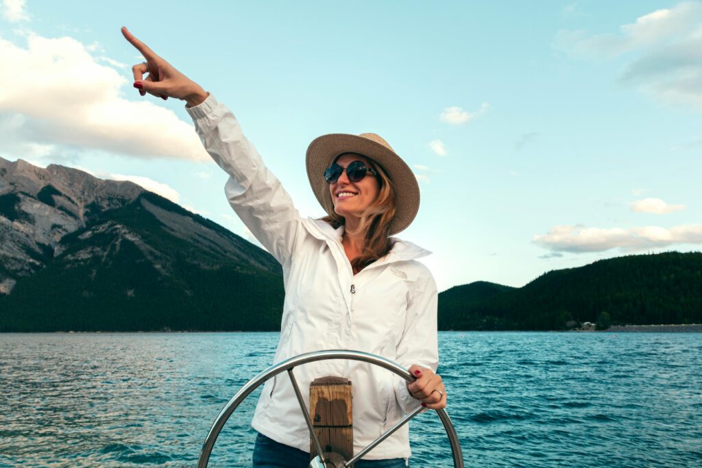 Smiling woman in sun hat and sunglasses pointing while steering a boat on Lake Minnewanka, Canada.