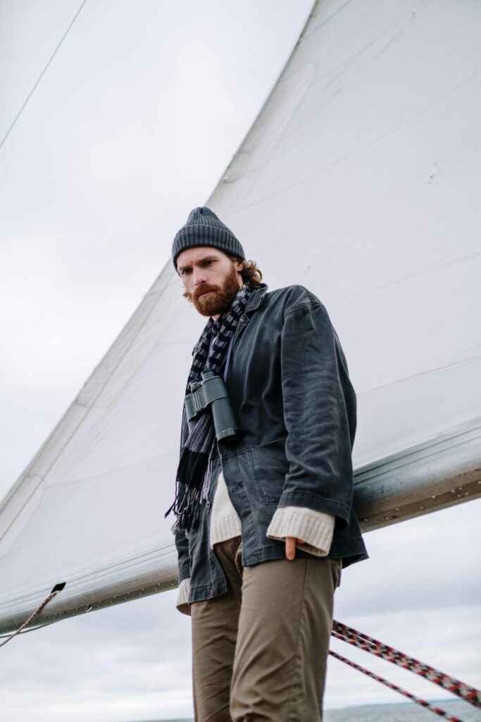 Bearded man wearing beanie and scarf standing on a sailboat under cloudy skies.